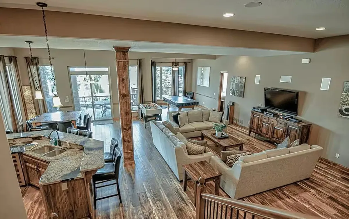 This image captures an integrated open-plan living, dining, and kitchen area. The cozy sofa arrangement faces the dining area and a kitchen bar, with large windows letting in abundant natural light. The space buzzes with potential for gatherings, from game nights to shared meals, all while enjoying views of the outside.