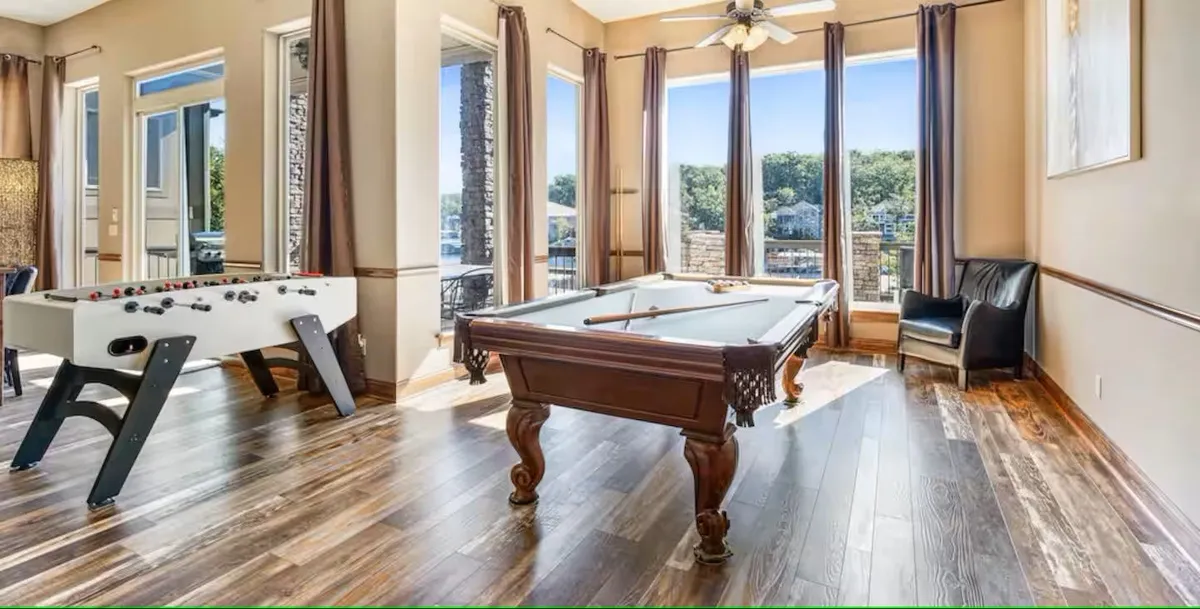 This image captures a lively game room scene featuring a pool table and foosball table as central attractions. Large windows bathe the space in natural light, while comfortable seating ensures everyone has a place to relax and socialize. The visual energy of friendly competition is palpable, and the views to the outdoors add openness to the oversized layout.
