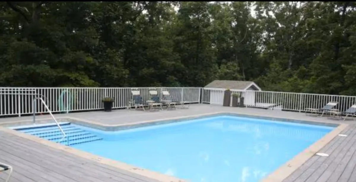 An inviting exterior scene showing a tranquil swimming pool surrounded by deck chairs and lush greenery. The area is designed for swimming, sunbathing, pool parties, and relaxed social gatherings in a resort-like setting.