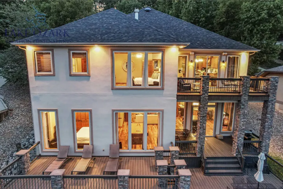 This image displays a spacious outdoor deck with comfortable seating, large windows showcasing the home's inviting interior, and stone pillars. The deck overlooks scenic surroundings and is perfectly set up for gatherings, dining, and enjoying sunset views.