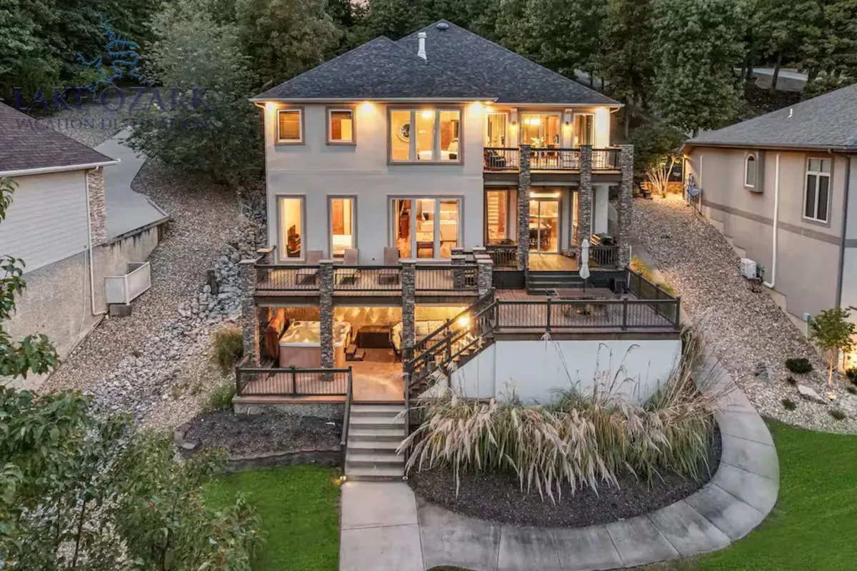 The image showcases the exterior of a luxurious single-family home at dusk, with expansive decks featuring railings, a spacious backyard, beautifully landscaped gardens, and glowing interior lights visible through large windows. The scene evokes a welcoming atmosphere perfect for gatherings, with sunset hues illuminating the deck and hints of cozy outdoor amenities like a fire pit.