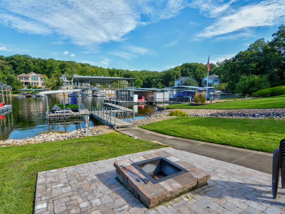 A spacious, green lakeside lawn with a fire pit, docks extending over the water, and boats moored nearby. The image highlights a welcoming area for evening campfires, fishing, or simply enjoying the lake atmosphere with family and friends.