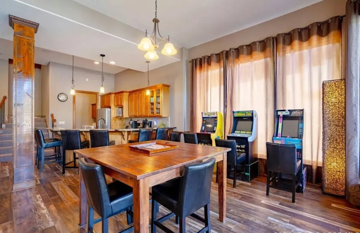A lively interior space featuring a dining table adjacent to arcade machines, set beside large windows and a connected kitchen area. The atmosphere is playful and warm, ideal for group meals, game nights, and casual gatherings.